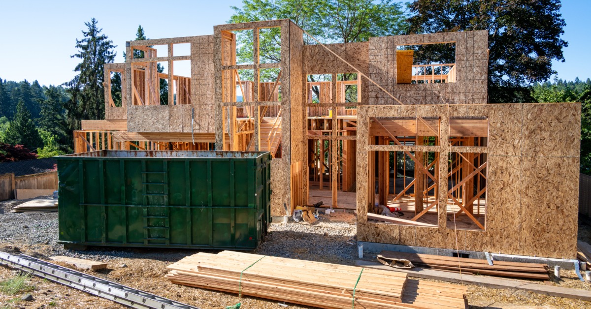 A two-story home under construction with exposed beams and wooden walls behind an empty large green dumpster.